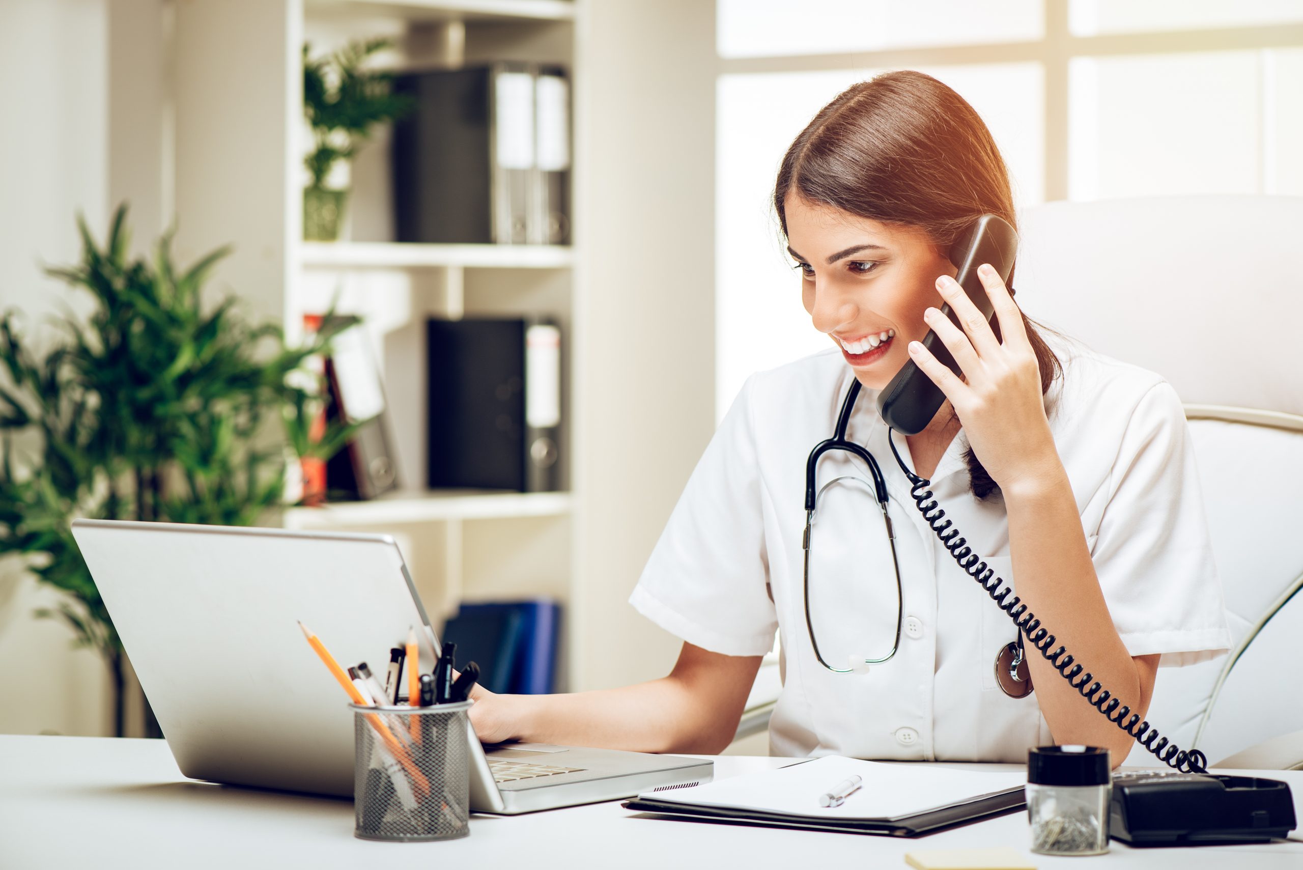 Beautiful young smiling female doctor sitting in her consulting room using phone and working on laptop.